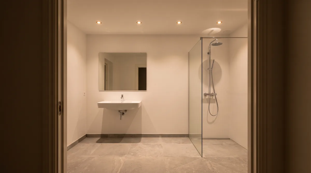 Badezimmer von der Tür aus gesehen mit vier eingeschalteten Einbaustrahlern in einer Reihe in der weißen Decke, Waschtisch mit Spiegel und einer Walk-in-Dusche mit Glaswand